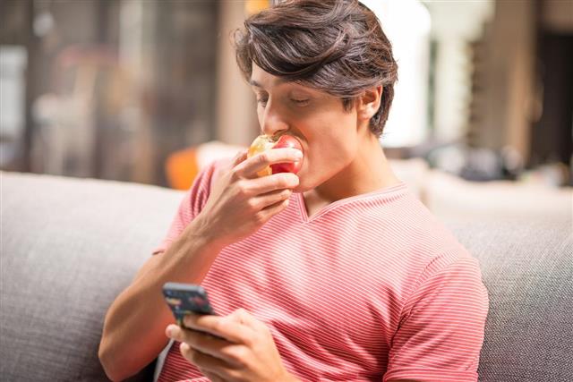 Man using smartphone and eating an apple on sofa
