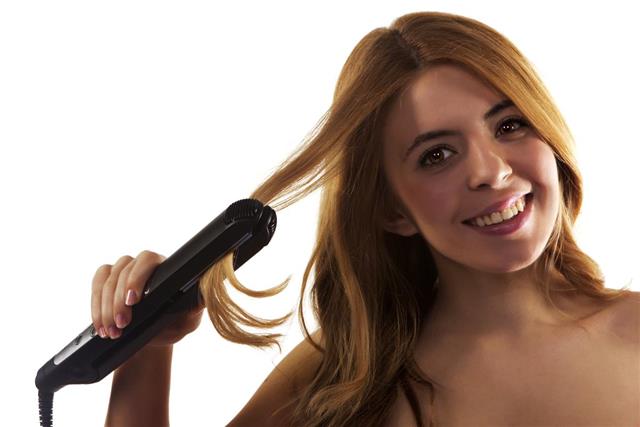 Beautiful young woman styling her hair with flat iron