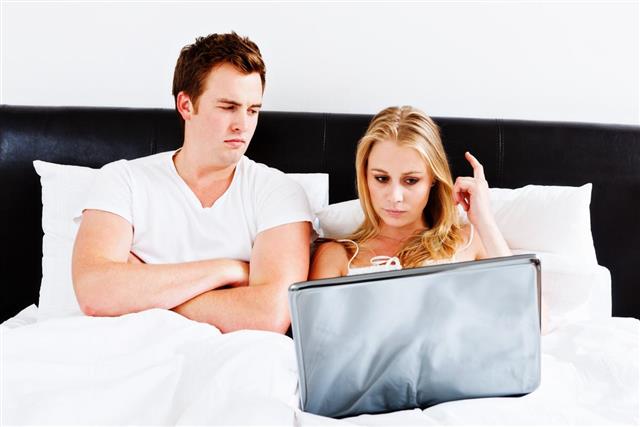 Young couple in bed: he sulks as she uses laptop