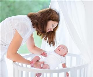 Young beautiful mother putting newborn baby into white round crib