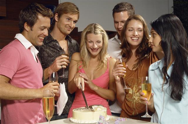 Young woman cutting birthday cake with friends holding champagne