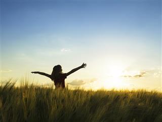 Girl stands in field looking at sunset with arms raised