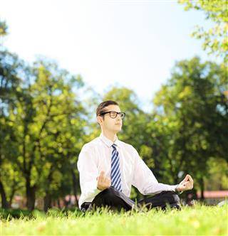 Young business person meditating seated on a grass in par