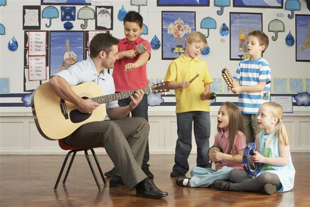 Male Teacher Playing Guitar With Pupils Having Music Lesson