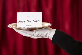 Butler holds 'Save the Date' notecard silver platter. Red curtains