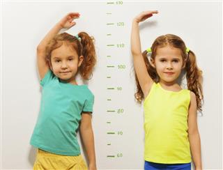 Two girls show height on wall scale at home