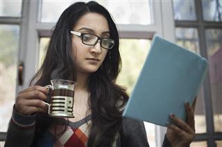 Young woman with coffee mug and book studying near window.